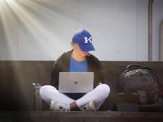 Student in blue UK baseball cap holds an open laptop while sitting on a bench.