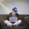 Student in blue UK baseball cap holds an open laptop while sitting on a bench.